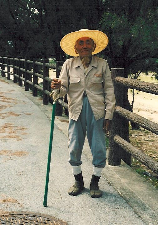 Elderly Okinawan farmer standing on a quiet path beside a wooden fence, wearing a wide straw hat, light jacket, and rolled-up trousers with gaiters. He leans on a green walking stick and smiles warmly, embodying resilience and grace.