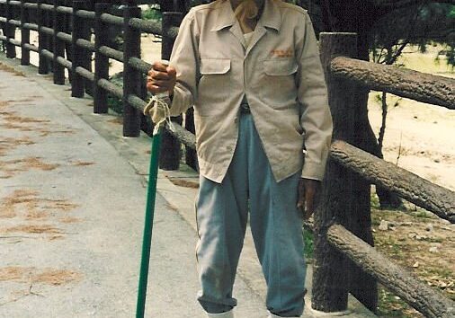 Elderly Okinawan farmer standing on a quiet path beside a wooden fence, wearing a wide straw hat, light jacket, and rolled-up trousers with gaiters. He leans on a green walking stick and smiles warmly, embodying resilience and grace.
